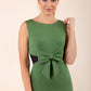 blonde model wearing Diva Catwalk Pencil sleeveless dress with rounded neckline and bow detail at the front with a contrasting black band in vineyard green front