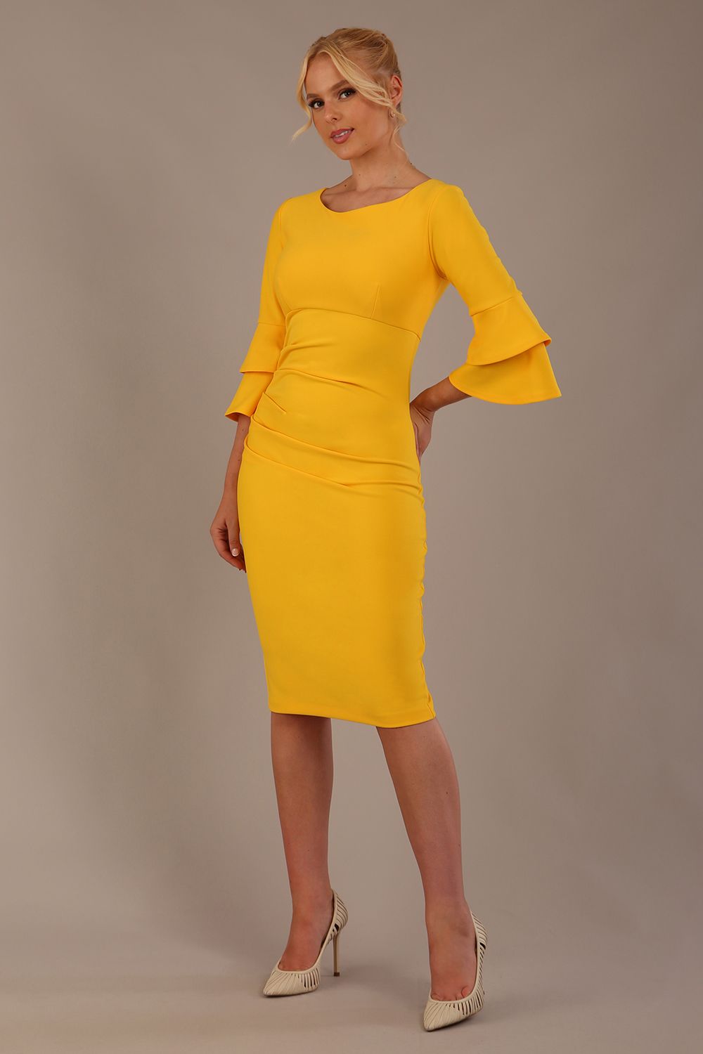 A woman wearing a sunshine yellow knee-length diva catwalk meridian pencil dress with a round neckline, double layered peplum three quarter length sleeves, and standing against a plain background.