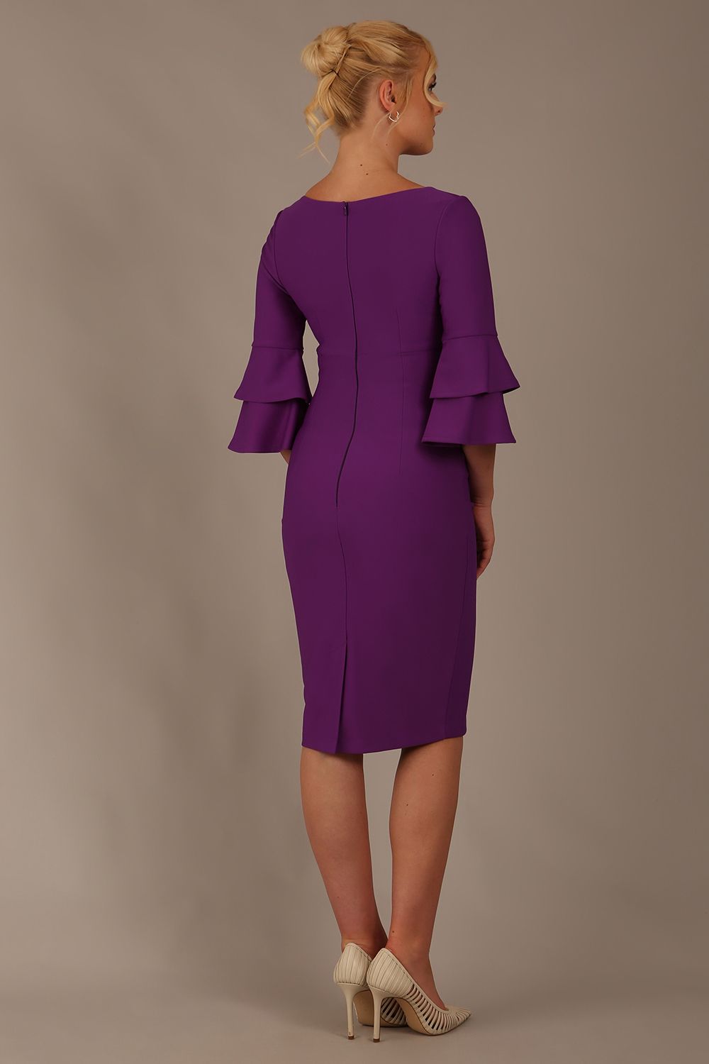 A woman wearing a royal purple knee-length diva catwalk meridian pencil dress with a round neckline, double layered peplum three quarter length sleeves, and standing against a plain background back view