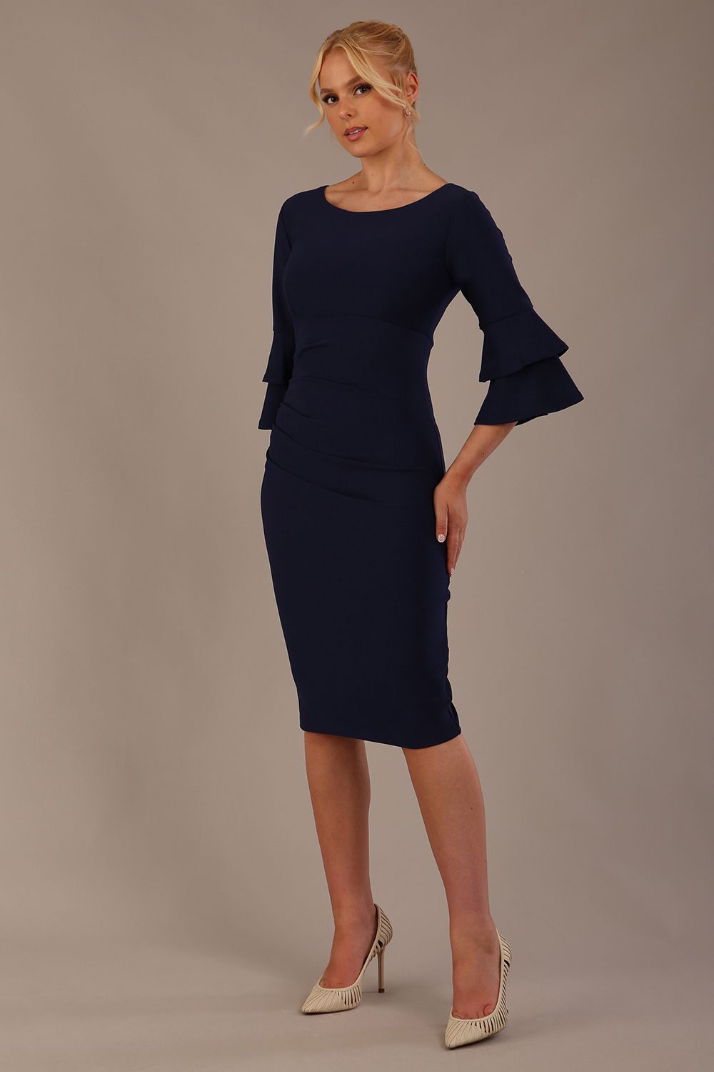 A woman wearing a navy blue knee-length diva catwalk meridian pencil dress with a round neckline, double layered peplum three quarter length sleeves, and standing against a plain background.