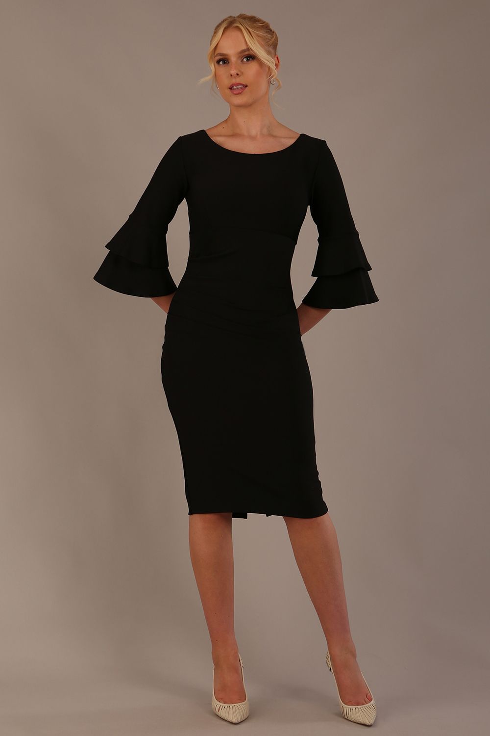 A woman wearing a black knee-length diva catwalk meridian pencil dress with a round neckline, double layered peplum three quarter length sleeves, and standing against a plain background.