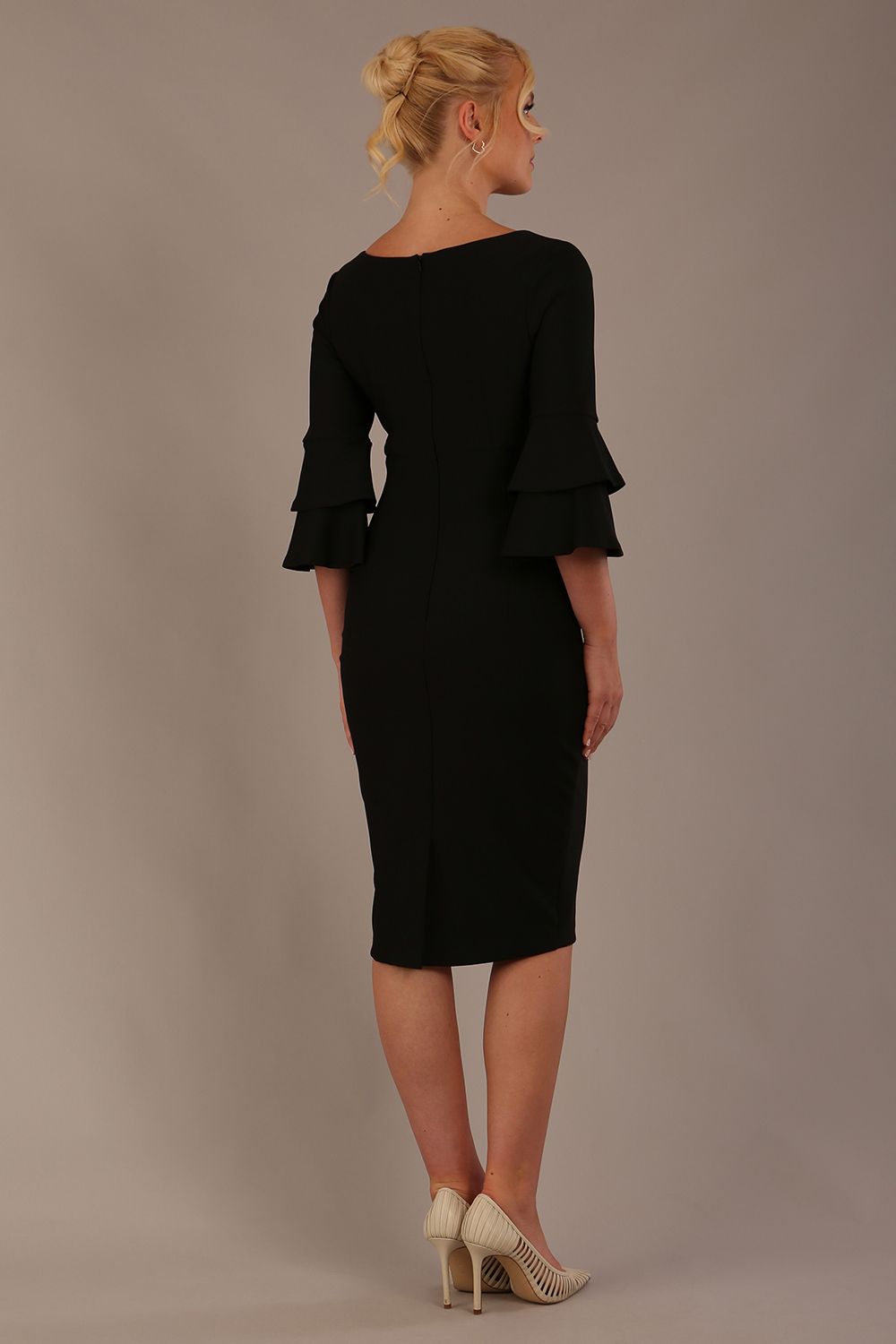 A woman wearing a black knee-length diva catwalk meridian pencil dress with a round neckline, double layered peplum three quarter length sleeves, and standing against a plain background back view