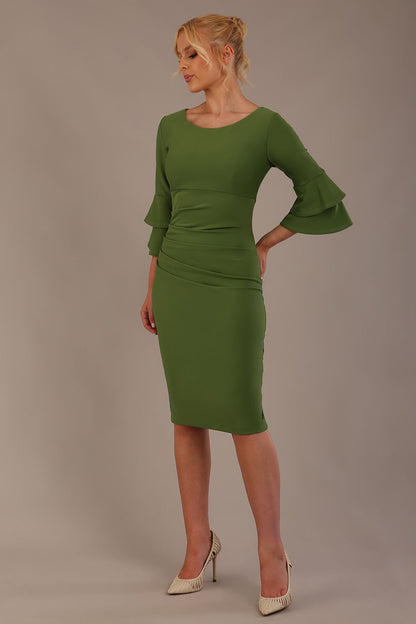 A woman wearing a vineyard green knee-length diva catwalk meridian pencil dress with a round neckline, double layered peplum three quarter length sleeves, and standing against a plain background.