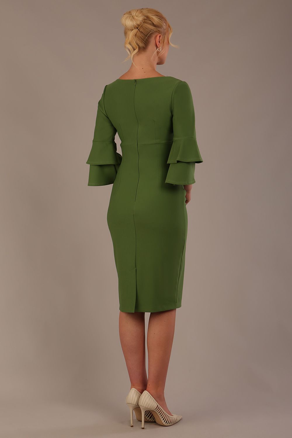 A woman wearing a vineyard green knee-length diva catwalk meridian pencil dress with a round neckline, double layered peplum three quarter length sleeves, and standing against a plain background back view