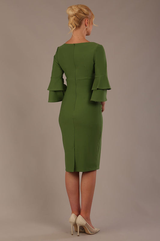 A woman wearing a vineyard green knee-length diva catwalk meridian pencil dress with a round neckline, double layered peplum three quarter length sleeves, and standing against a plain background back view