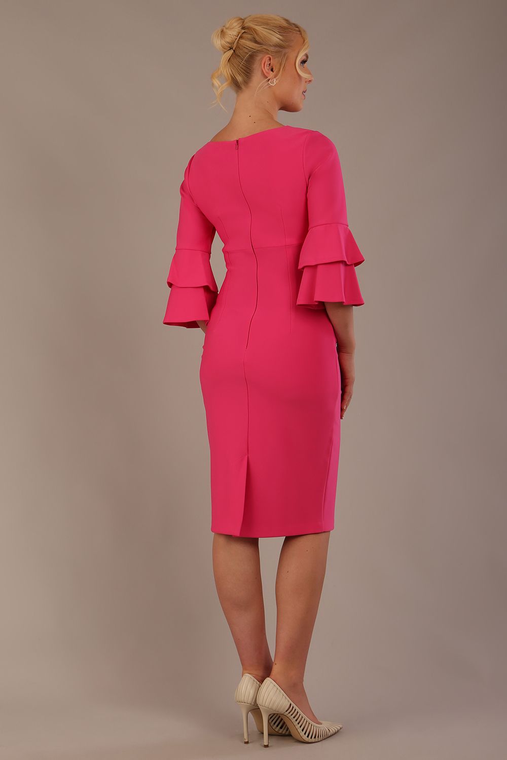 A woman wearing a hibiscus pink knee-length diva catwalk meridian pencil dress with a round neckline, double layered peplum three quarter length sleeves, and standing against a plain background back view