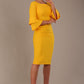 A woman wearing a sunshine yellow knee-length diva catwalk meridian pencil dress with a round neckline, double layered peplum three quarter length sleeves, and standing against a plain background.
