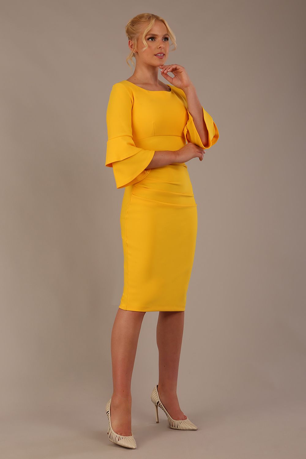 A woman wearing a sunshine yellow knee-length diva catwalk meridian pencil dress with a round neckline, double layered peplum three quarter length sleeves, and standing against a plain background.