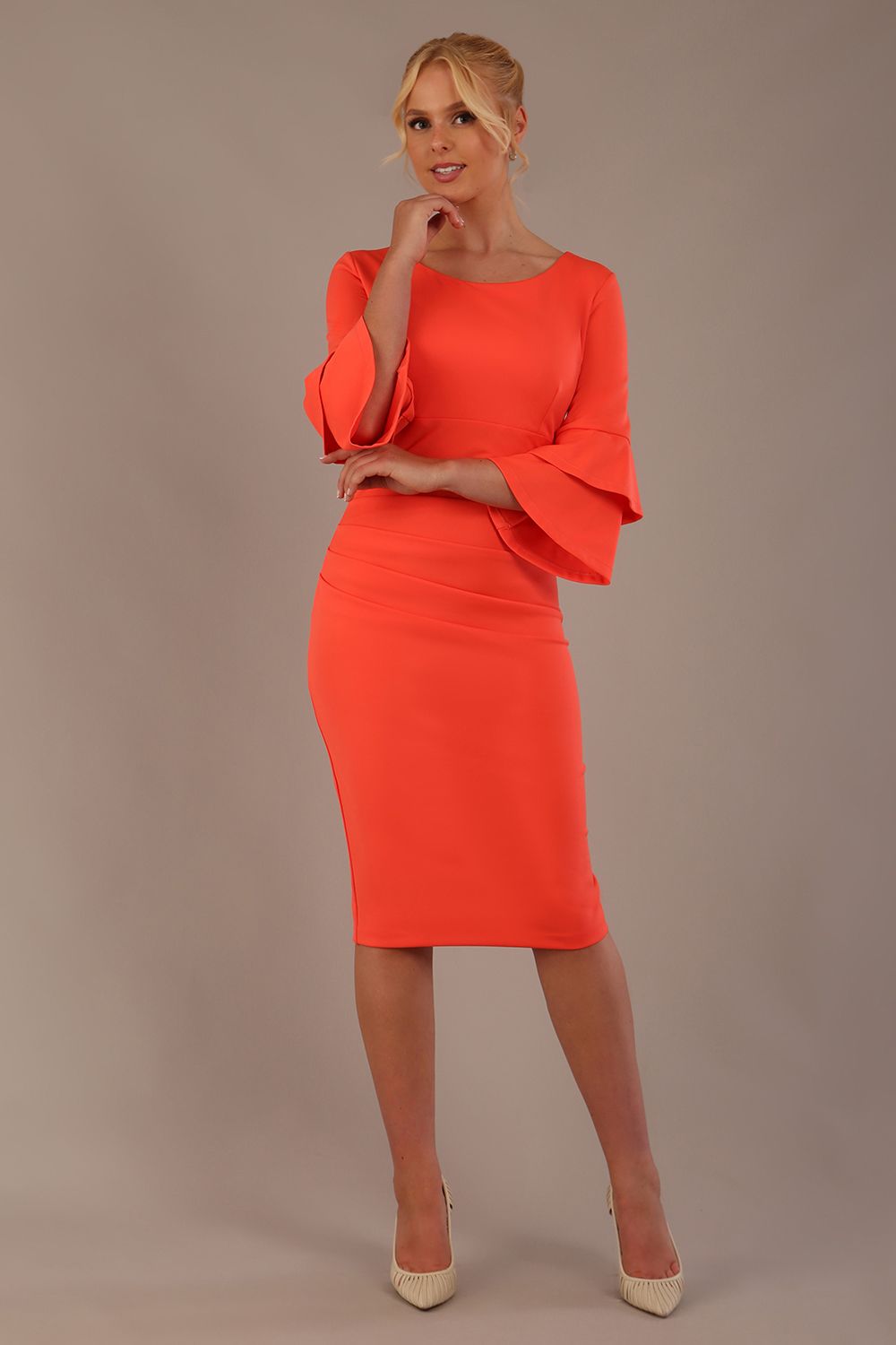 A woman wearing a hot coral knee-length diva catwalk meridian pencil dress with a round neckline, double layered peplum three quarter length sleeves, and standing against a plain background.