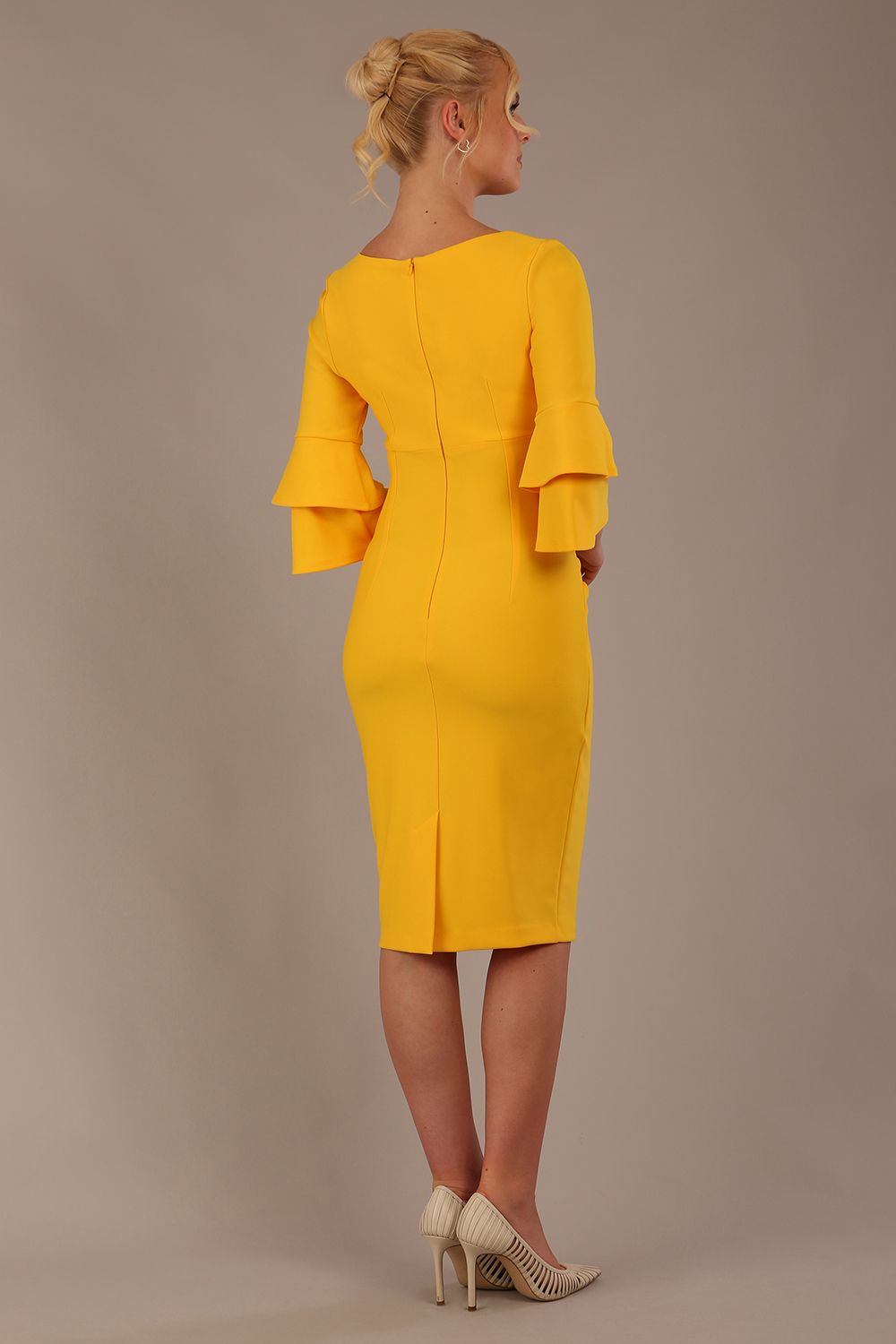 A woman wearing a sunshine yellow knee-length diva catwalk meridian pencil dress with a round neckline, double layered peplum three quarter length sleeves, and standing against a plain background back view