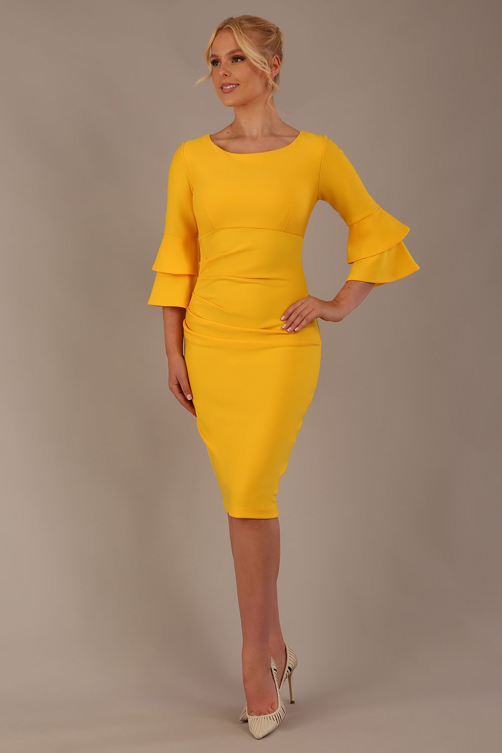 A woman wearing a sunshine yellow knee-length diva catwalk meridian pencil dress with a round neckline, double layered peplum three quarter length sleeves, and standing against a plain background
