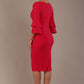 A woman wearing a yarrow pink knee-length diva catwalk meridian pencil dress with a round neckline, double layered peplum three quarter length sleeves, and standing against a plain background back side view