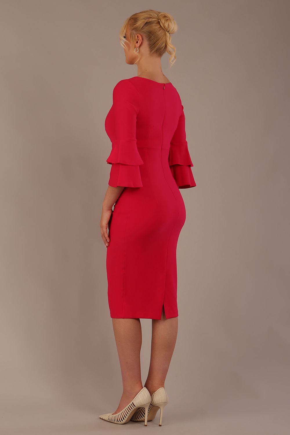 A woman wearing a yarrow pink knee-length diva catwalk meridian pencil dress with a round neckline, double layered peplum three quarter length sleeves, and standing against a plain background back side view