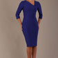 A woman modeling a palace blue diva catwalk castleford pencil dress with an asymmetric neckline, bow waistband, and pleated skirt