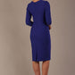 A woman modeling a palace blue diva catwalk castleford pencil dress with an asymmetric neckline, bow waistband, and pleated skirt back view
