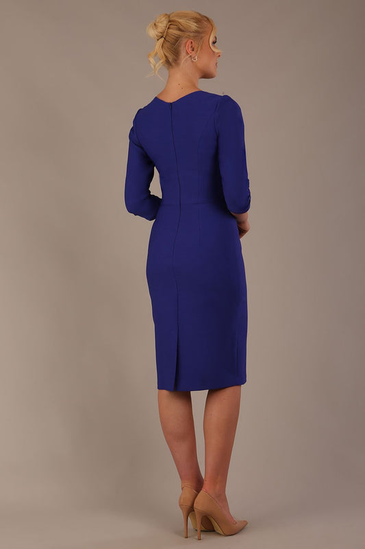 A woman modeling a palace blue diva catwalk castleford pencil dress with an asymmetric neckline, bow waistband, and pleated skirt back view