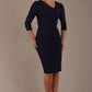 A woman modeling a navy blue diva catwalk castleford pencil dress with an asymmetric neckline, bow waistband, and pleated skirt