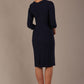 A woman modeling a navy blue diva catwalk castleford pencil dress with an asymmetric neckline, bow waistband, and pleated skirt back view