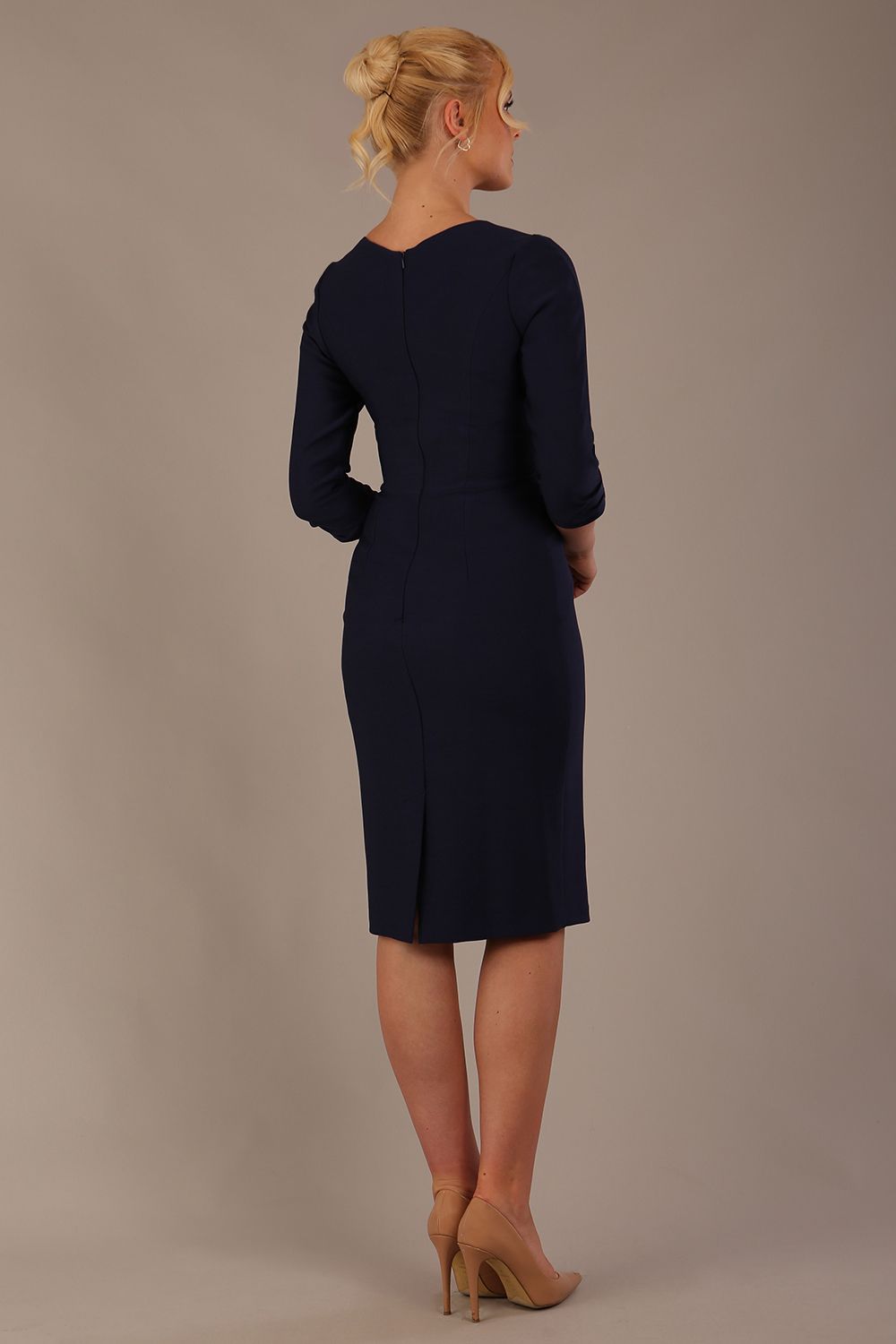 A woman modeling a navy blue diva catwalk castleford pencil dress with an asymmetric neckline, bow waistband, and pleated skirt back view