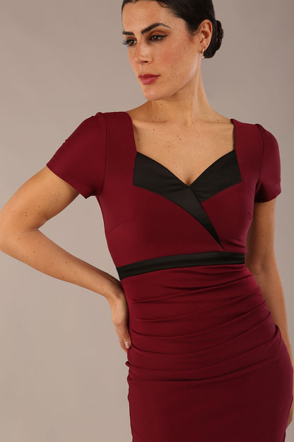 model is wearing a diva catwalk dress in burgundy with black satin detail contrast on the neckline and waistband short sleeves and pleating details with knee length