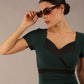 model is wearing a diva catwalk dress in forest green with black satin detail contrast on the neckline and waistband short sleeves and pleating details with knee-length