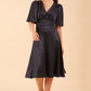 brunette model wearing diva catwalk cambrian satin swing dress with bell short sleeve wide asymmetric waistband and pleating under bust and low v-neckline in navy front