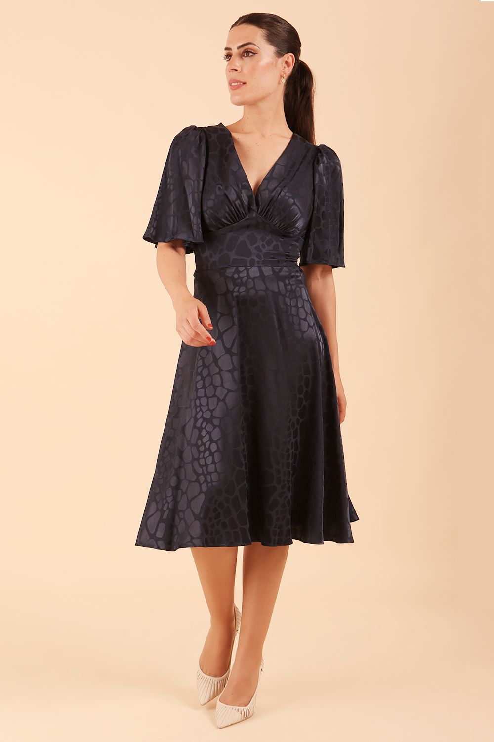 brunette model wearing diva catwalk cambrian satin swing dress with bell short sleeve wide asymmetric waistband and pleating under bust and low v-neckline in navy front