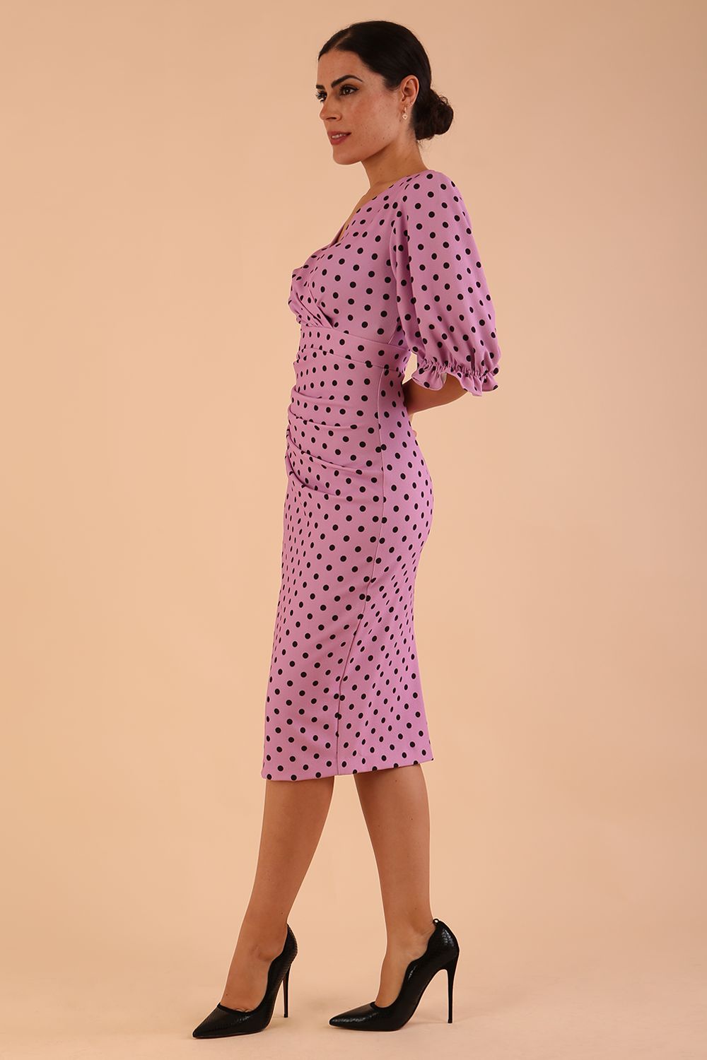 brunette model wearing diva catwalk palacio pencil fitted dress with three quarter puffed sleeve and pleating across the body with overlapping v-neckline in pink polka dot front side
