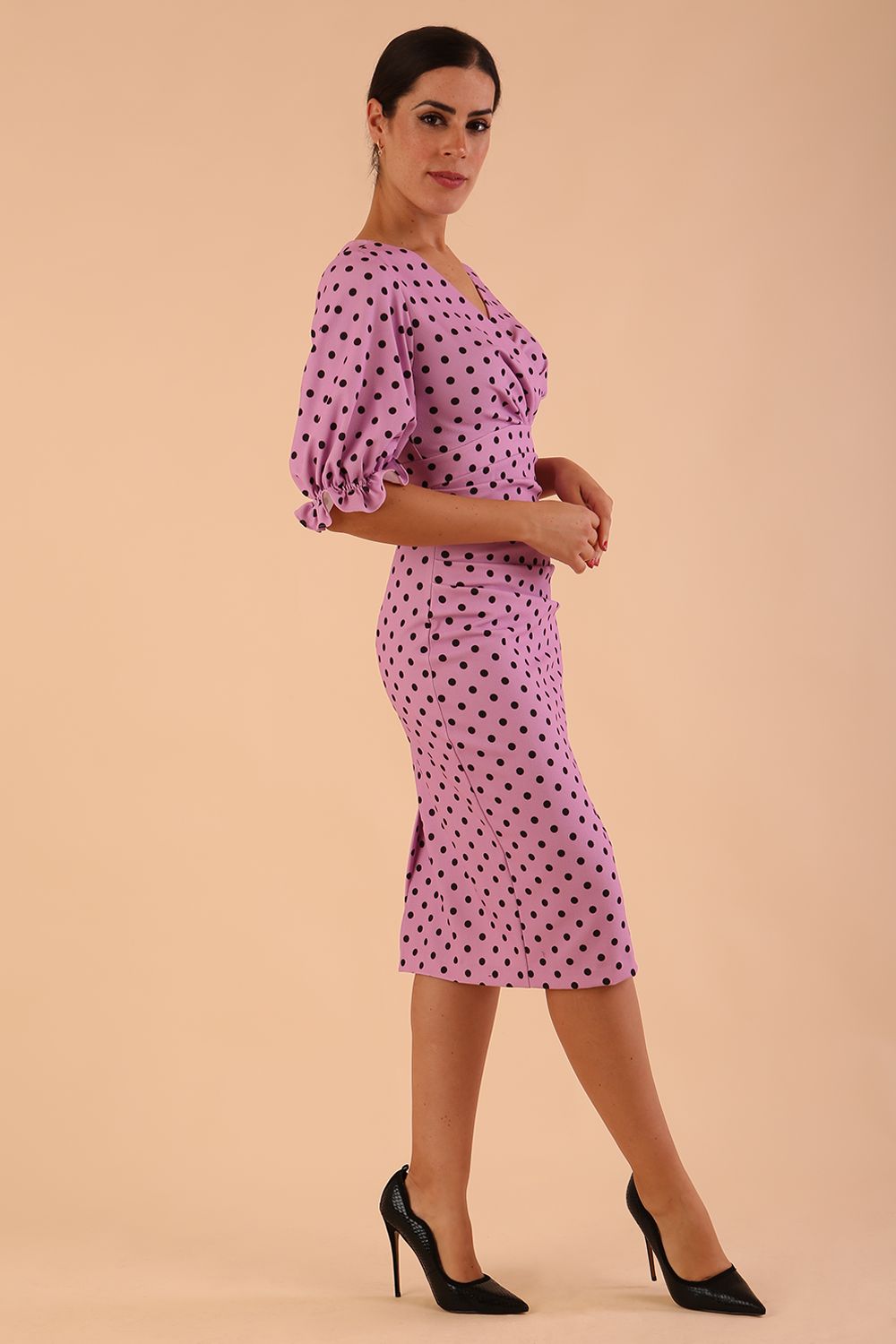 brunette model wearing diva catwalk palacio pencil fitted dress with three quarter puffed sleeve and pleating across the body with overlapping v-neckline in pink polka dot front side