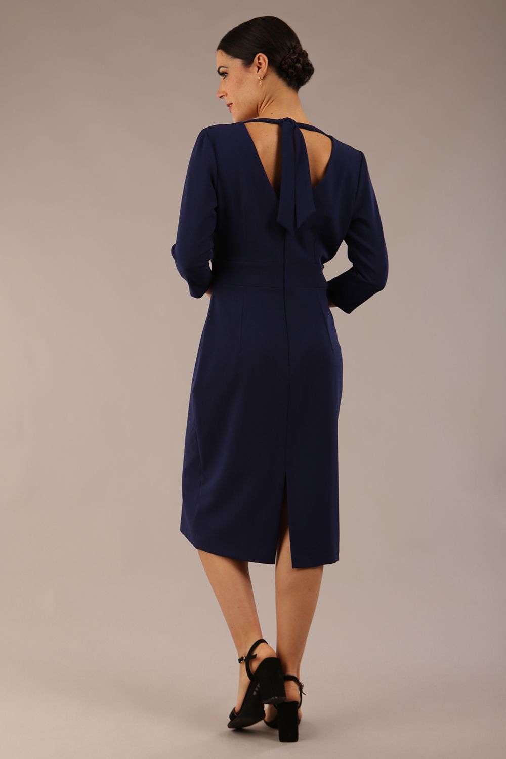 brunette model wearing diva catwalk wimpole a-line flattering dress with lowered v-neck and three quarter sleeve and cinching waistband and widened skirt with pleating in navy blue back