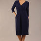 brunette model wearing diva catwalk wimpole a-line flattering dress with lowered v-neck and three quarter sleeve and cinching waistband and widened skirt with pleating in navy blue front