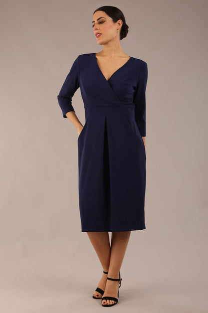 brunette model wearing diva catwalk wimpole a-line flattering dress with lowered v-neck and three quarter sleeve and cinching waistband and widened skirt with pleating in navy blue front