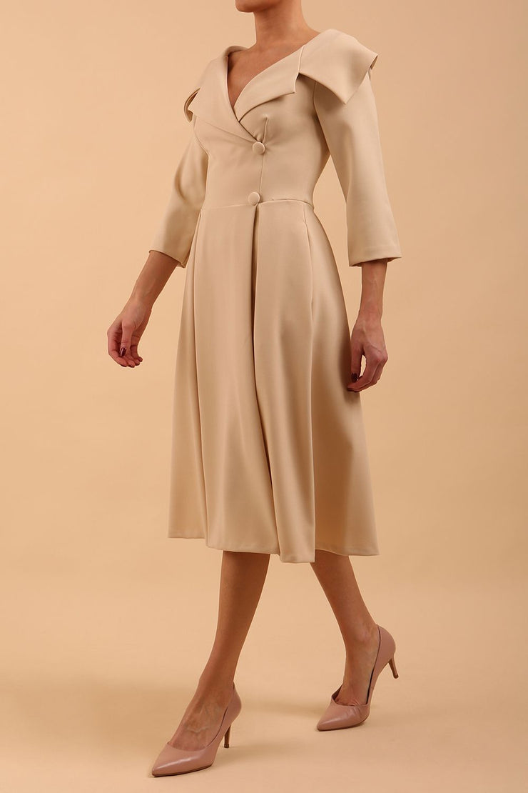 Brunette model is wearing a sleeved sandshell beige oversized collar swing dress with button detail at the front and pockets in the skirt