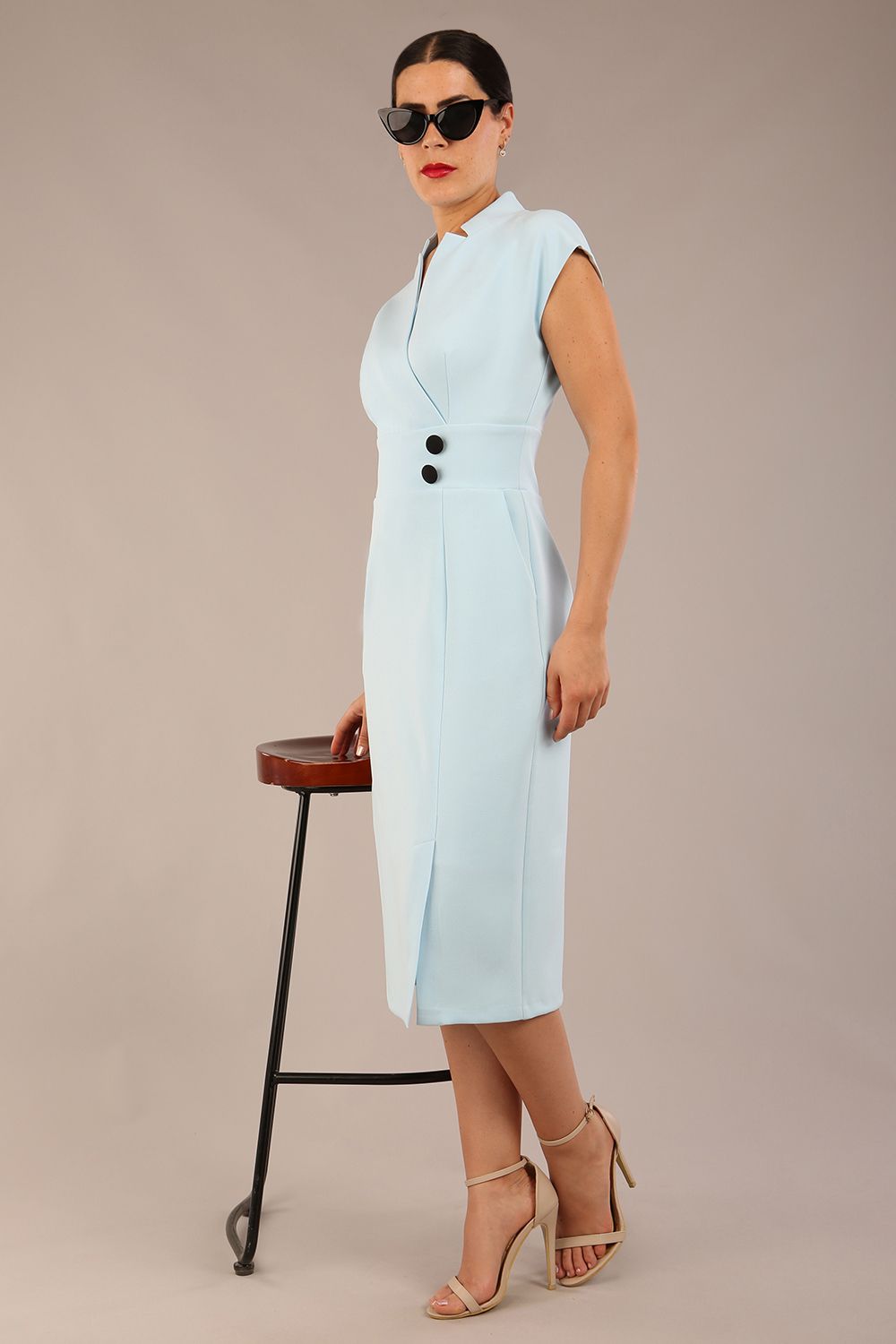 brunette Model is wearing a diva catwalk Derby pencil skirt dress cap sleeves with buttons details at the waistband and star neckline below the knee length with pockets in sky blue colour front image with sunglasses with a chair