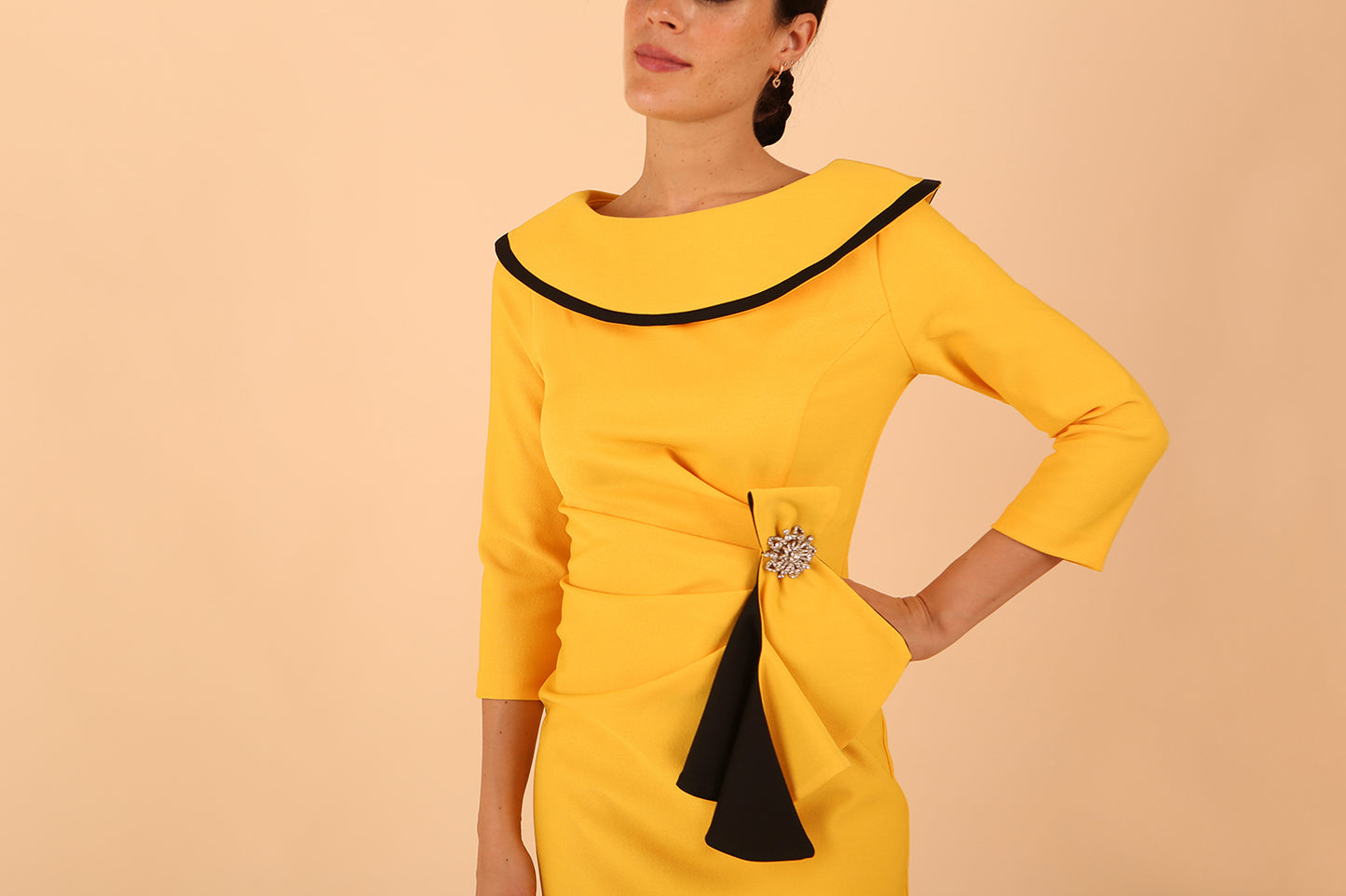 odel wearing diva catwalk Seed Elsye 3/4 Sleeve Knee Length Pencil Dress with Fold over bateau collar neckline with contrast piping and Bow feature on the left side of the bodice with decorative brooch in Daffodil Yellow close up colour front