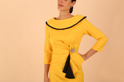 odel wearing diva catwalk Seed Elsye 3/4 Sleeve Knee Length Pencil Dress with Fold over bateau collar neckline with contrast piping and Bow feature on the left side of the bodice with decorative brooch in Daffodil Yellow close up colour front