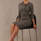 The model is wearing a diva catwalk Juliette 3/4 Sleeve Knee Length dress with bow detail in castlerock grey colour, sitting on a chair against a plain background