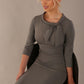 The model is wearing a diva catwalk Juliette 3/4 Sleeve Knee Length dress with bow detail in castlerock grey colour, sitting on a chair against a plain background
