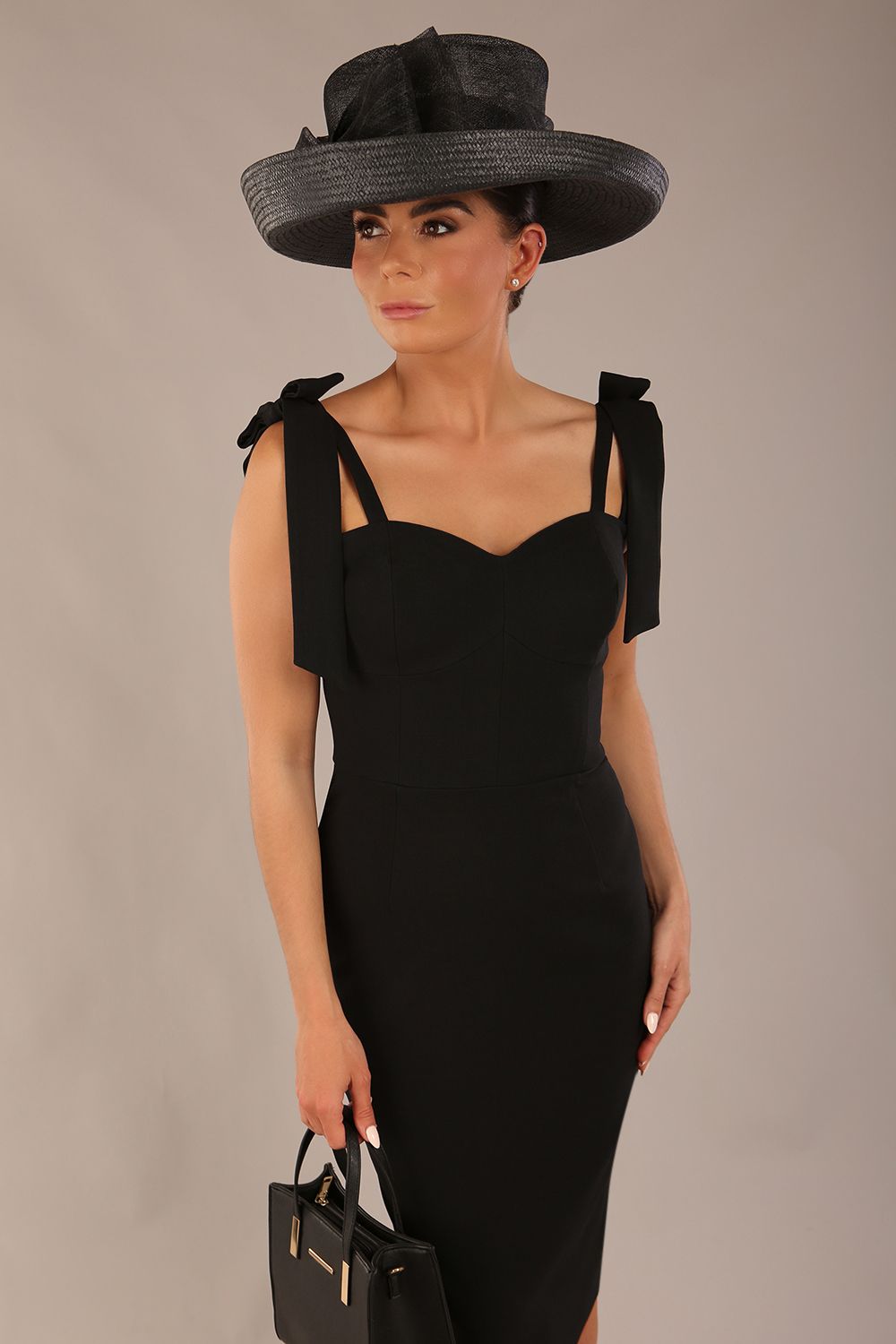 brunette model wearing a diva catwalk Philippa Pencil Wiggle Dress sleeveless style, tie detail on the shoulders knee length and sweetheart neckline in black colour front with hat