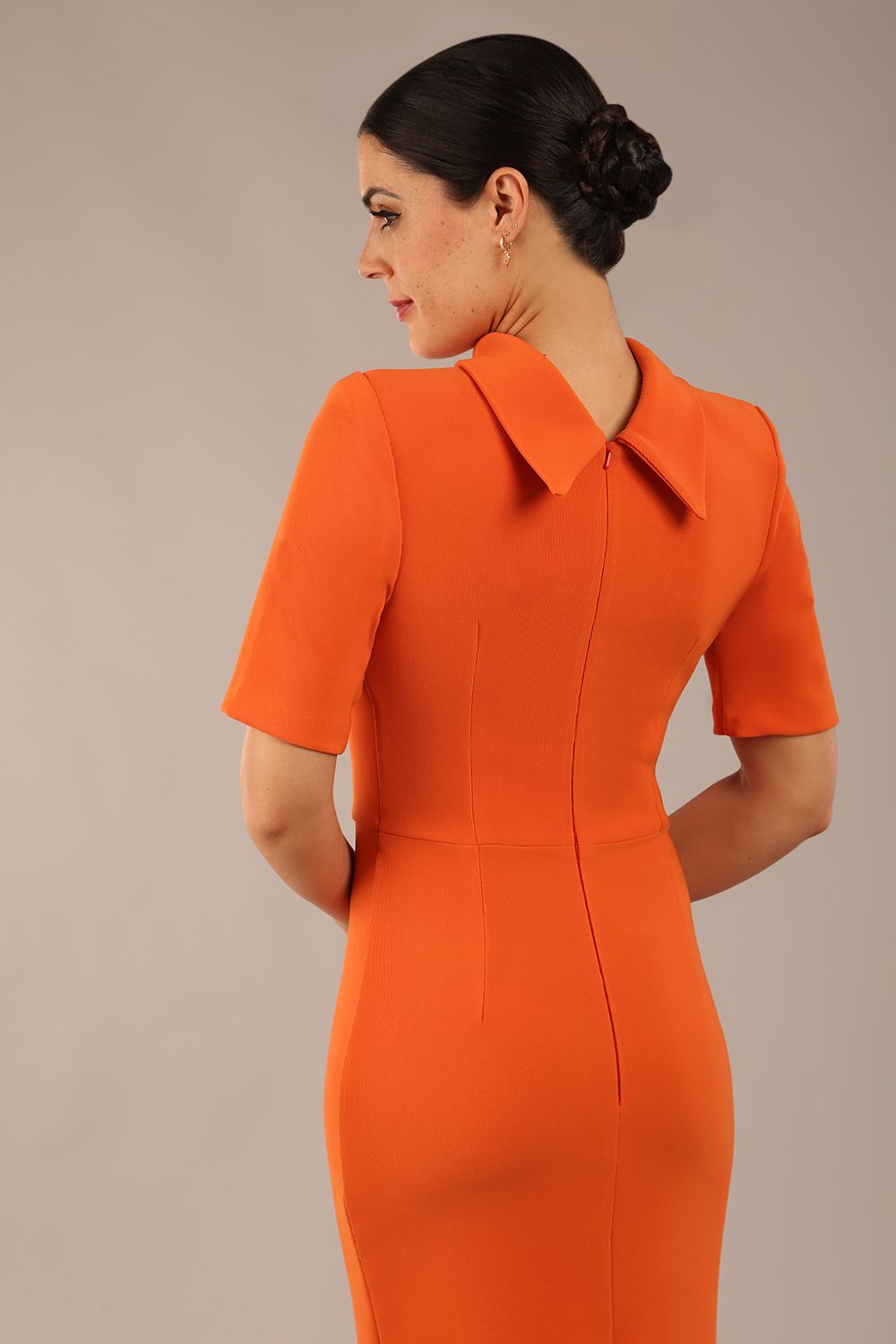 brunette model is wearing a diva catwalk Highmoor Short Sleeved Pencil Dress high neckline and shoulders pad in amber orange colour back image