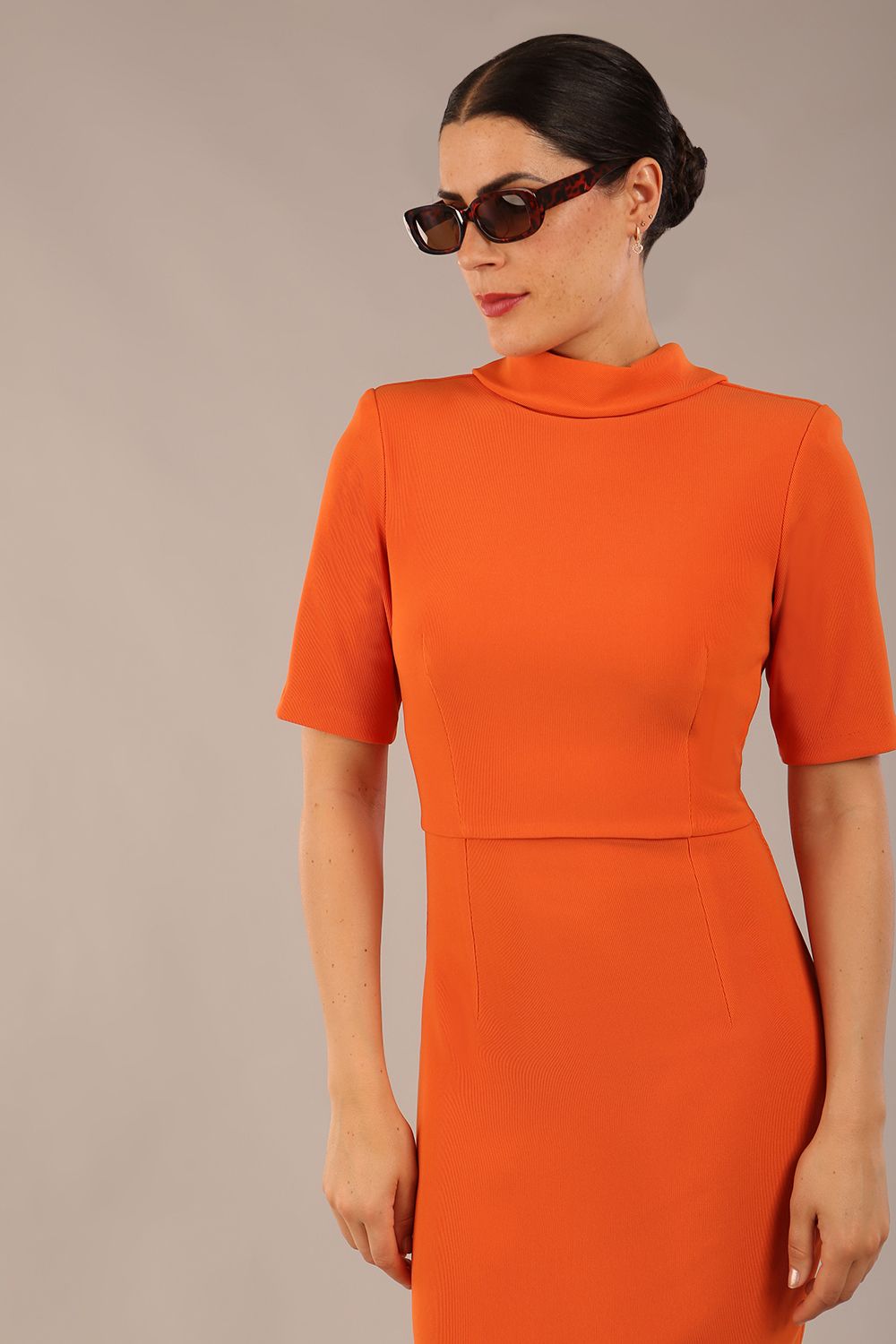 brunette model is wearing a diva catwalk Highmoor Short Sleeved Pencil Dress high neckline and shoulders pad in amber orange colour front image