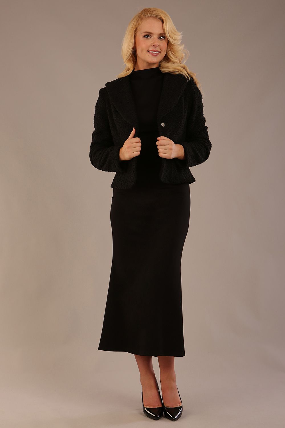 Woman wearing a black outfit with a textured jacket and the Kaden high neck midaxi dress in jet black, showing the sleek silhouette and cap sleeves against a plain background 