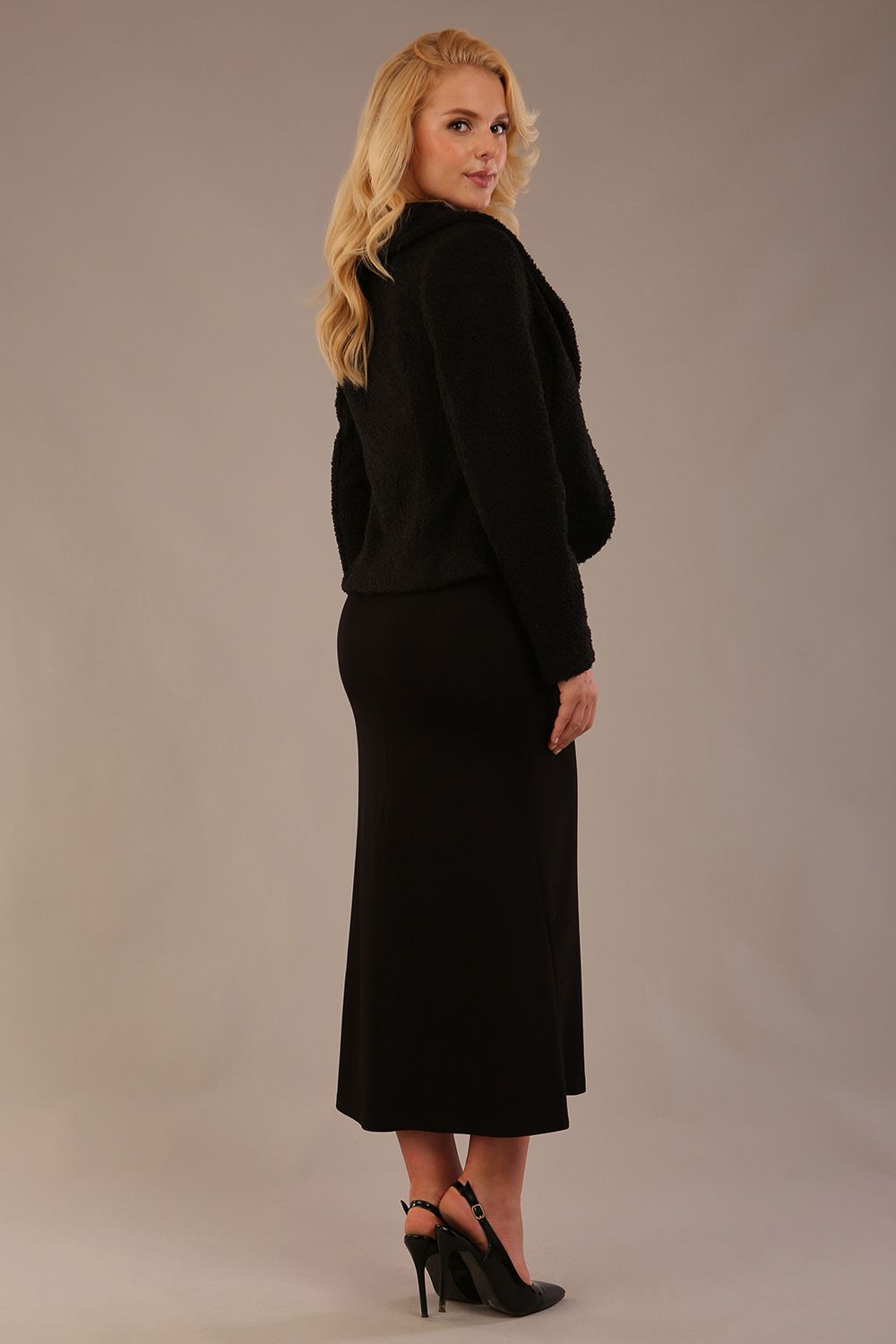 Woman wearing a black outfit with a textured jacket and the Kaden high neck midaxi dress in jet black, showing the sleek silhouette and cap sleeves against a plain background back view