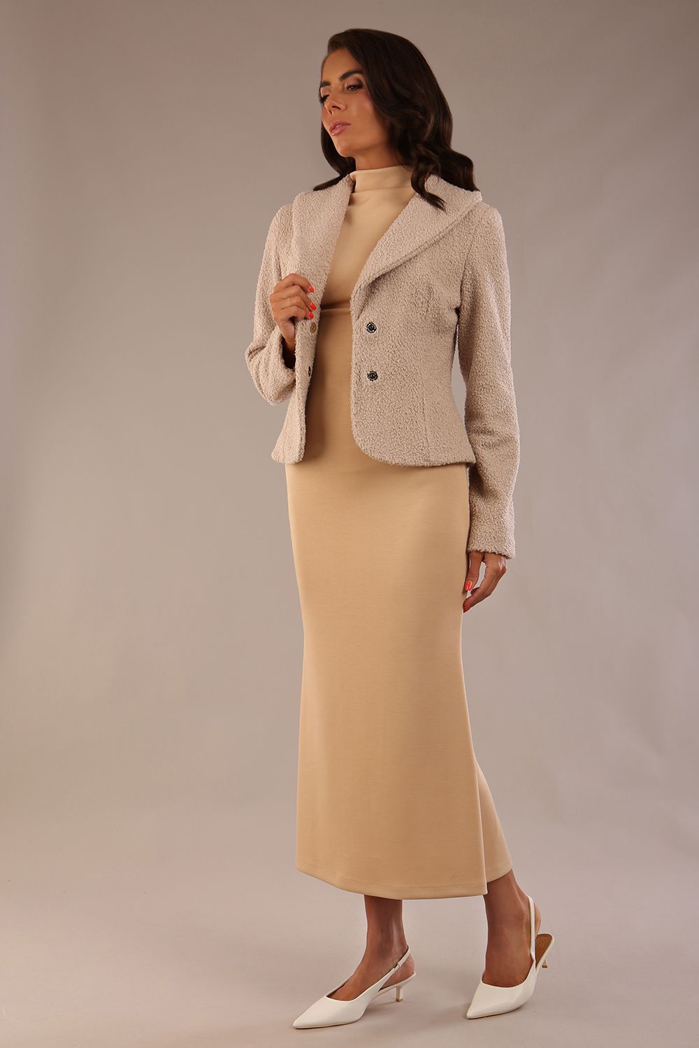 Woman wearing a beige outfit with a textured jacket and the Kaden high neck midaxi dress in cafe au lait cream, showing the sleek silhouette and cap sleeves against a plain background 