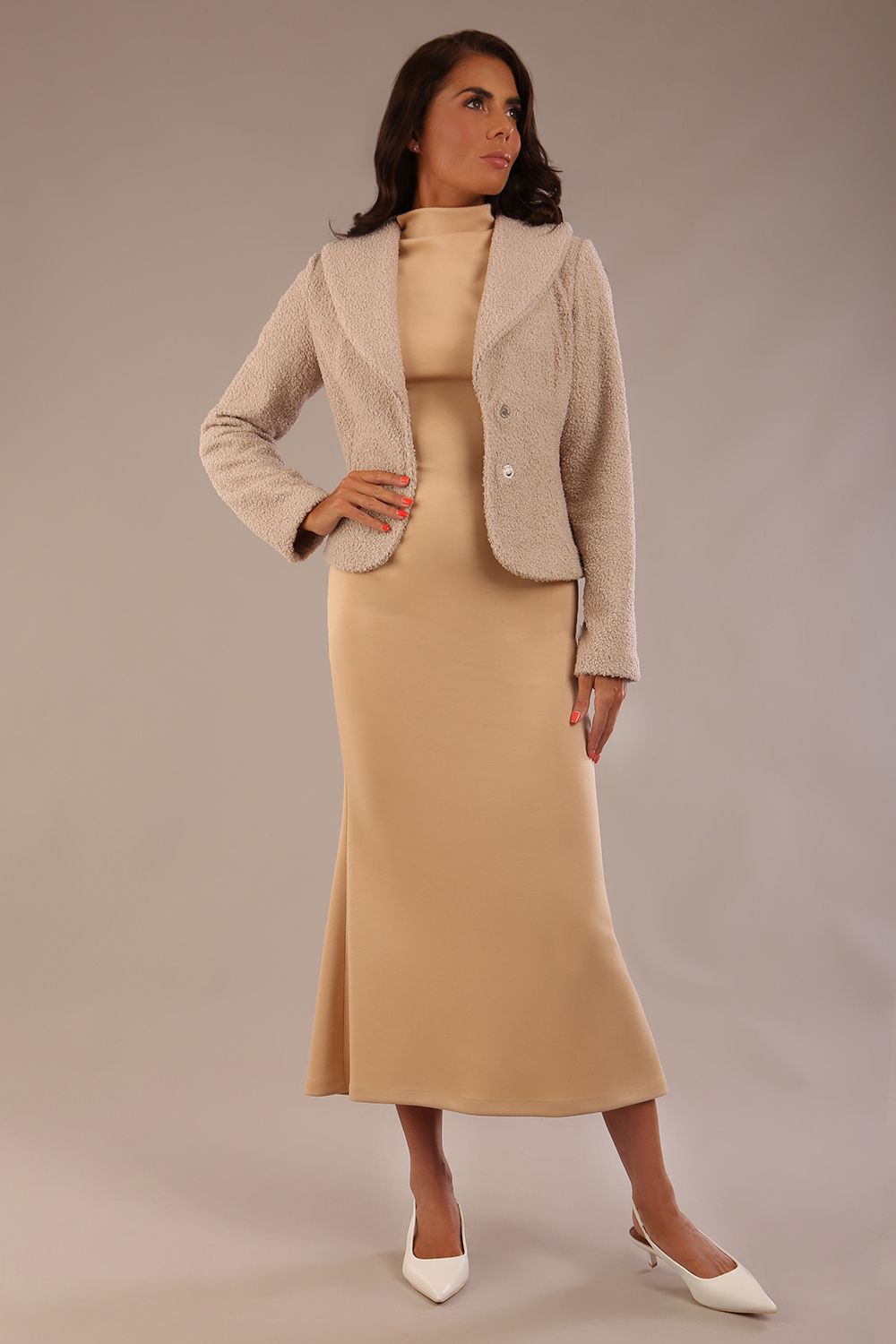 Woman wearing a beige outfit with a textured jacket and the Kaden high neck midaxi dress in cafe au lait cream, showing the sleek silhouette and cap sleeves against a plain background 