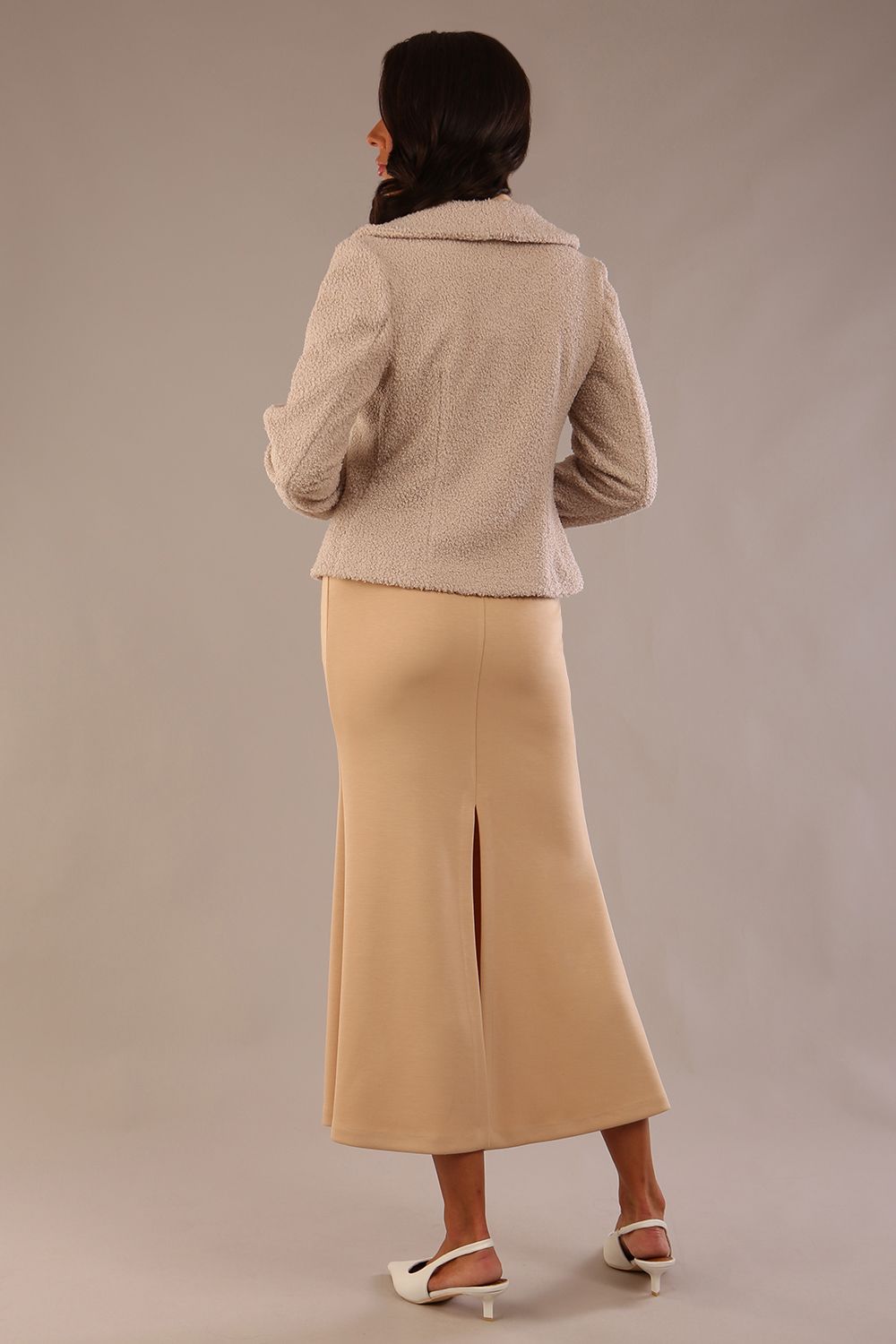 Woman wearing a beige outfit with a textured jacket and the Kaden high neck midaxi dress in cafe au lait cream, showing the sleek silhouette and cap sleeves against a plain background  back view