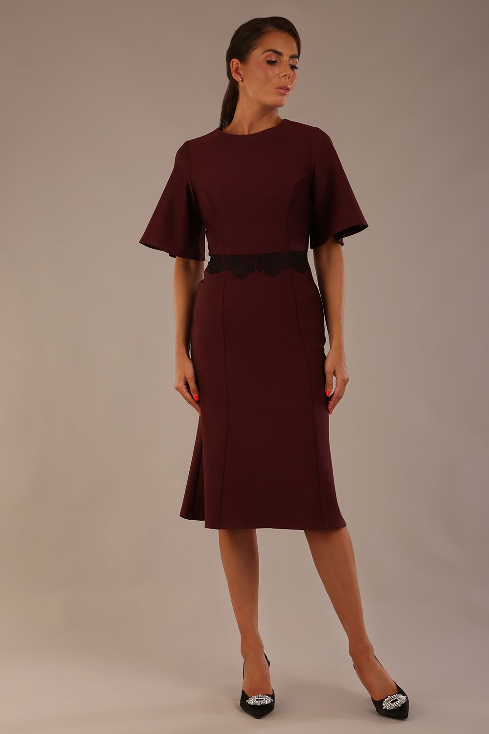 Model wearing the Lovell dress in port royale, showcasing the fluted sleeves and black lace waistband against a beige background