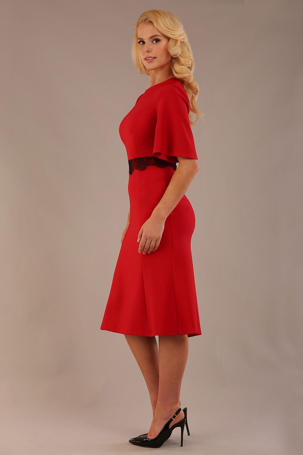 Model wearing the Lovell dress in salsa red, showcasing the fluted sleeves and black lace waistband against a beige background