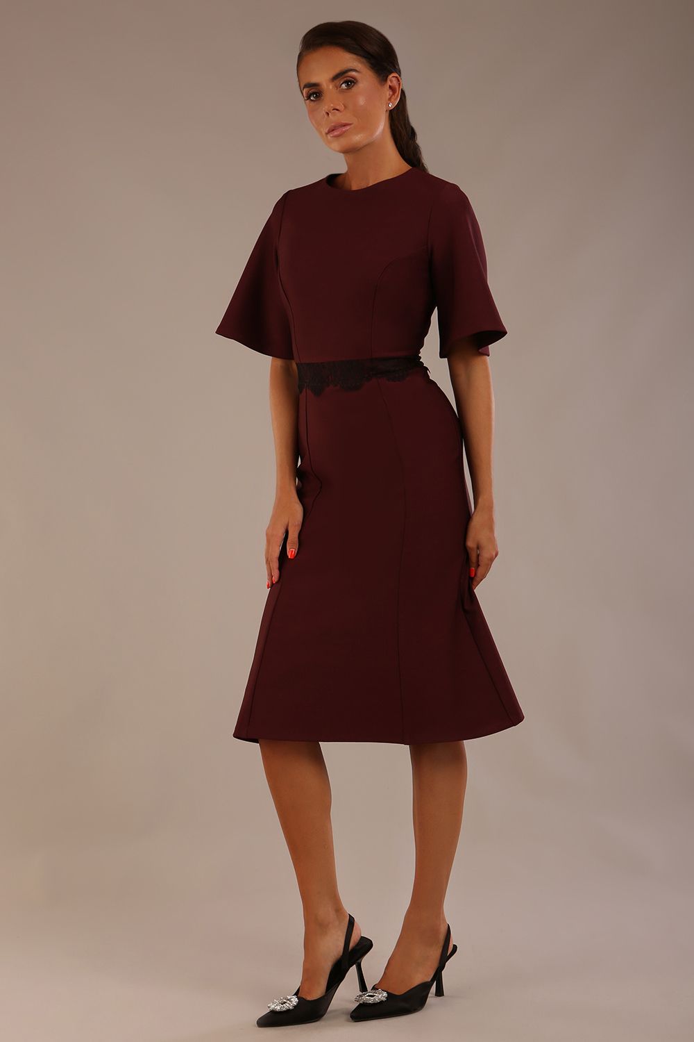 Model wearing the Lovell dress in port royale, showcasing the fluted sleeves and black lace waistband against a beige background