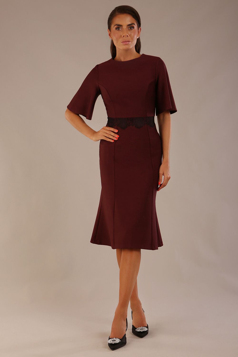 Model wearing the Lovell dress in port royale, showcasing the fluted sleeves and black lace waistband against a beige background